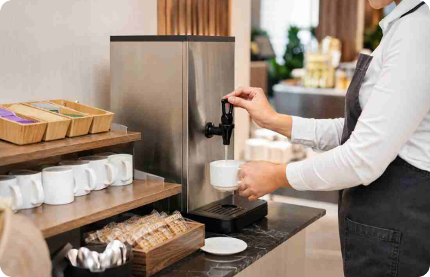 Staff dispensing hot water from a stainless steel Burco tower boiler into a white mug at a tea and coffee station