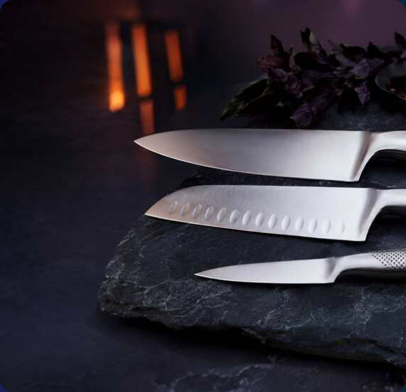 Three Global stainless steel knives — chef’s knife, santoku and utility — on a dark slate surface