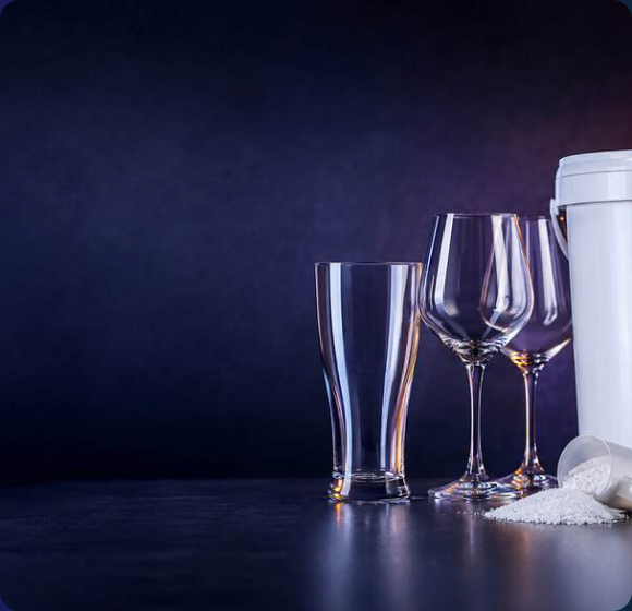 Clear beer and wine glasses beside a tub of Renovate glasswash powder with a scoop on a dark surface